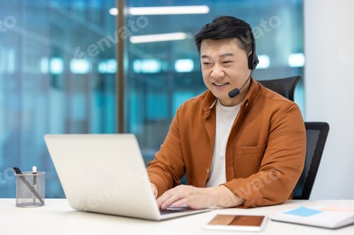 Preview: Asian businessman working at modern office using a headset while smiling at laptop with a