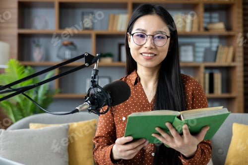 Preview: Asian woman recording podcast at home with microphone and book, smiling at camera, expressing