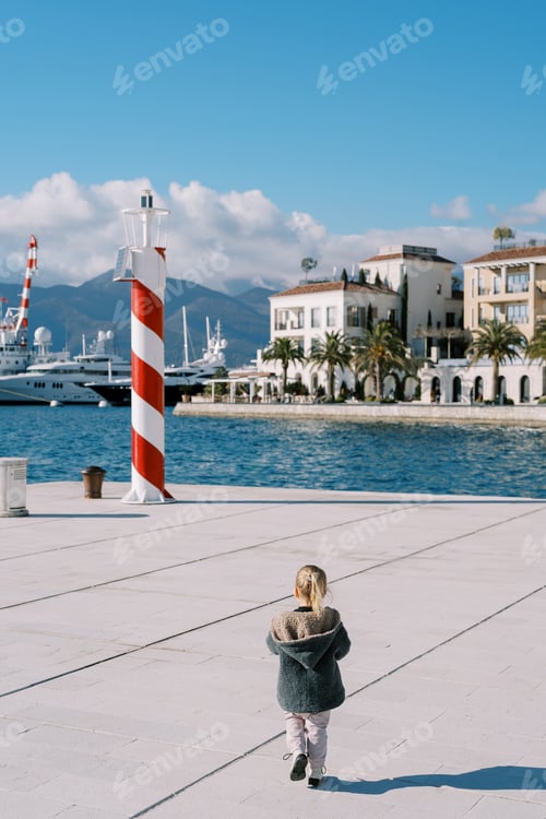 Preview: Little girl walks along the pier towards the striped lighthouse. Back view