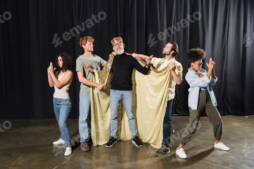 Preview: bearded art director standing in costume of king near multicultural students playing guards on