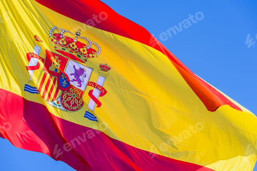 Preview: Red and yellow Spain flag with royal shield waving in the wind isolated against blue sky background.
