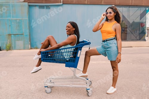 Preview: Playful young two multiracial women posing with shopping cart in urban street