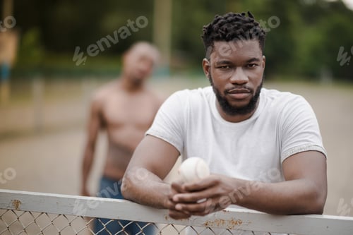 Preview: young handsome african american man with baseball ball leaning on fence on court