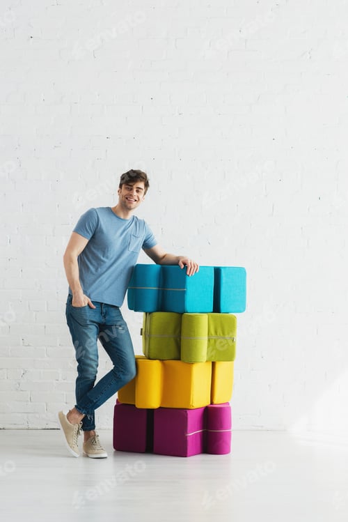 Preview: cheerful man standing and smiling near colorful poufs near brick wall