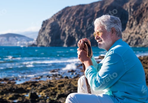 Preview: Senior man sitting at sunset light on the beach holding a walking stick because of body aches