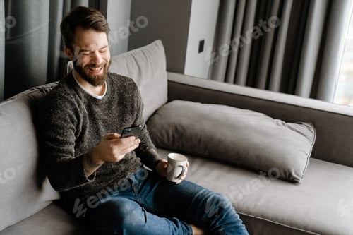 Preview: Bearded man using mobile phone and drinking coffee while sitting on sofa
