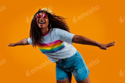 Preview: Young woman wearing rainbow t-shirt and funny hat is posing with open arms and orange background