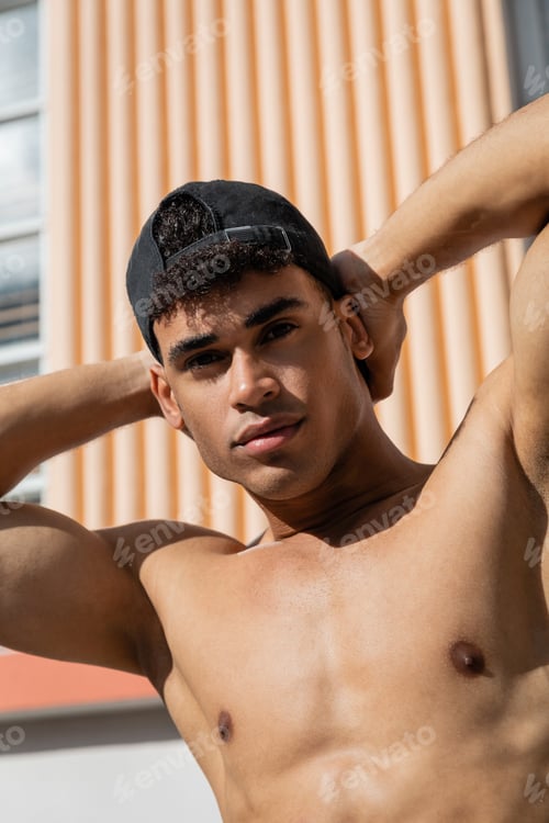 Preview: Portrait of sexy and muscular young cuban man in baseball cap touching neck and looking at camera