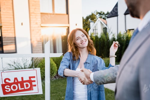 Preview: Happy redhead woman with keys shaking hands with broker near sign with sold lettering on blurred