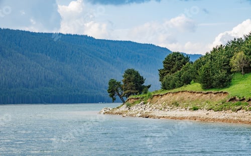 Preview: Deciduous trees grow on ledge above lake against backdrop of mountain range covered with vegetation