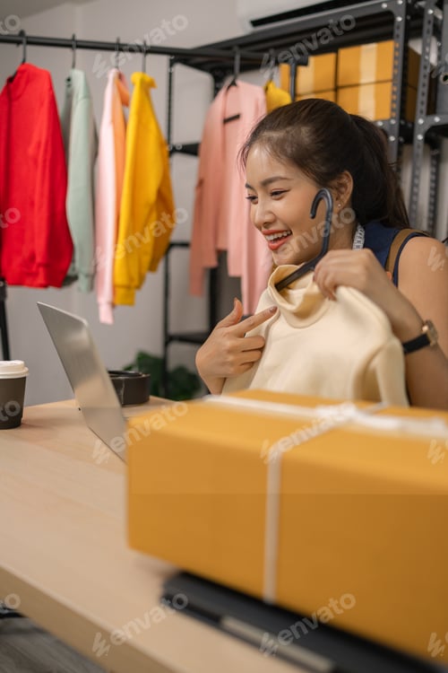 Preview: Young female entrepreneur smiling while livestreaming to sell clothes from her home office.