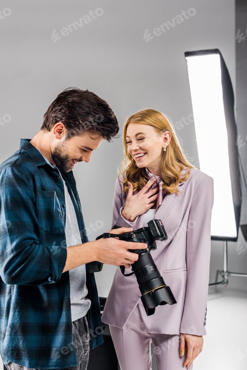Preview: handsome young photographer and smiling female model using photo camera and laughing in studio