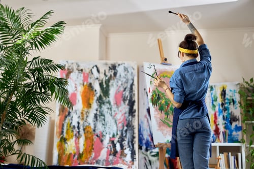 Preview: Back view of female artist raised her hand with paintbrush in it while creating a