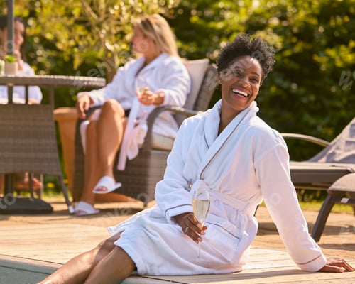 Preview: Mature Woman Wearing Robe Sitting With Feet In Pool Drinking Champagne On Spa Day