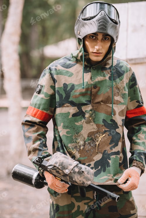 Preview: young male paintball player in camouflage covered by paintball splash outdoors