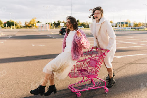 Preview: Gorgeous girl in long skirt sitting in pink shopping cart. Outdoor photo of two relaxed women at pa