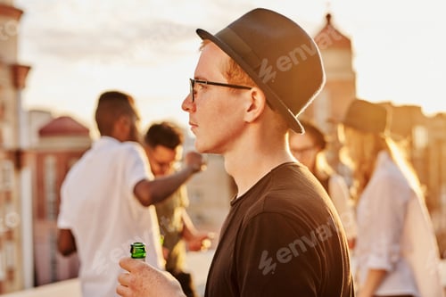 Preview: Young Man in Hat Standing on Rooftop with Beer Bottle