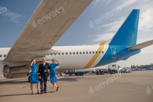 Preview: Full length shot of happy pilot walking together with two stewardesses in bright blue uniform