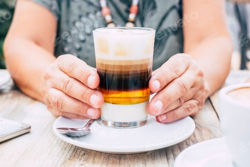 Preview: Close up with aged female hands holding a cup of multi coloured coffee for breakfast at the bar