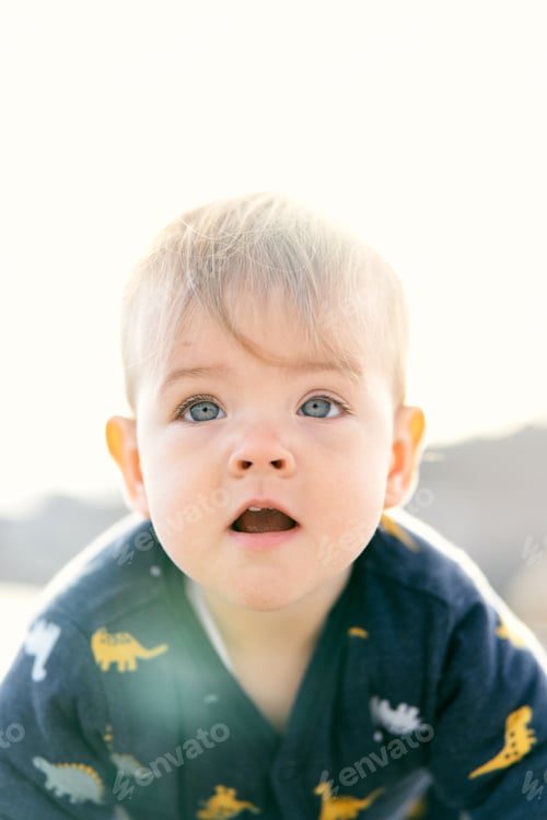 Preview: Surprised toddler with open mouth in overalls sits on a pebble beach. Close-up. Portrait
