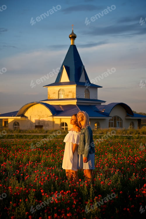 Preview: Two beautiful girls in summer dresses standing on the poppy field beside the orthodox church