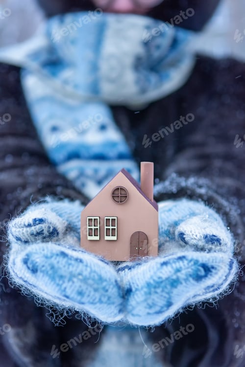 Preview: a woman in fluffy mittens holds a small house in her hands on a cold winter day.