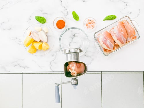 Preview: Manual meat grinder with glass bowl,boneless chicken thighs,peeled onions,garlic on a marble table .