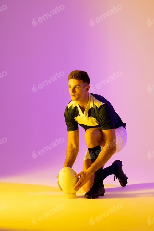 Preview: Caucasian male rugby player crouching with rugby ball over pink lighting