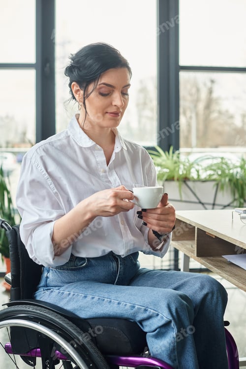 Preview: dedicated woman with mobility disability in wheelchair holding cup of coffee while in office