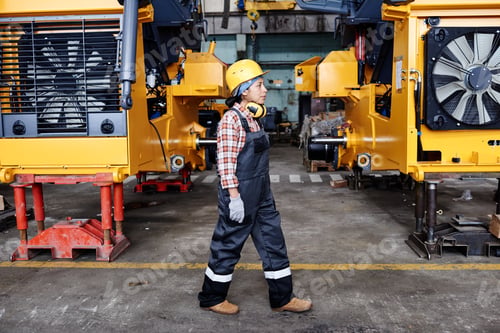 Preview: Young female technician in overalls, harhat and gloves moving along machines