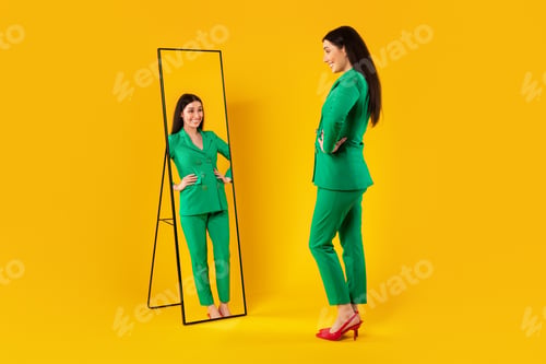 Preview: Happy lady wearing long jacket trouser suit and looking at her reflection in mirror, elegant posing