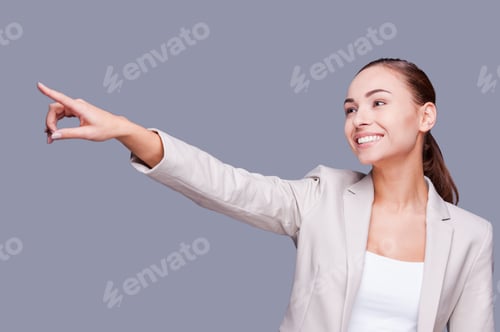 Preview: Look at that! Smiling young businesswoman pointing away while standing against grey background