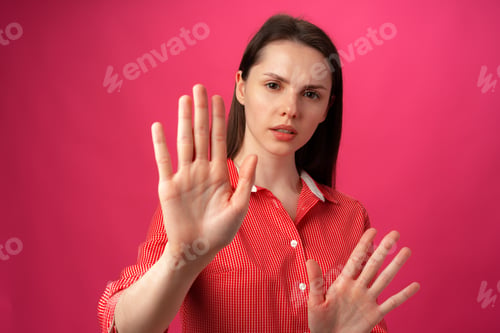Preview: Young brunette woman does stop sign with palm against pink background