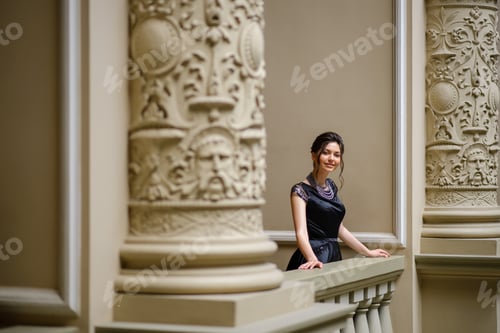 Preview: Young beautiful woman in a black dress peeps through the columns.
