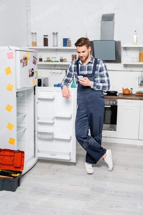 Preview: full length of smiling handyman texting on smartphone while leaning on fridge in kitchen