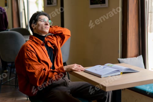 Preview: a young guy in glasses sits in a cafe with a book and looks thoughtfully out window