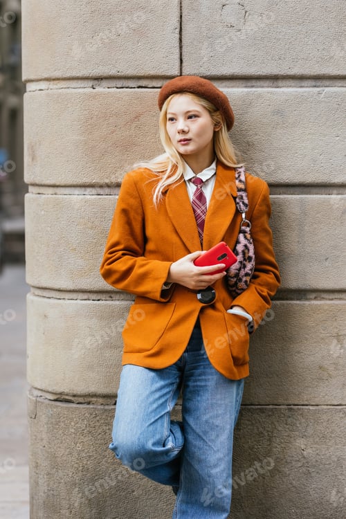 Preview: Stylish Asian woman holding mobile phone while leaning on a wall outdoors.