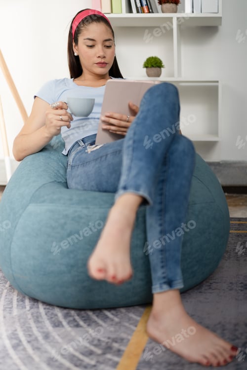 Preview: Young woman, concentrated, reading on a digital tablet and drinking a cup of coffee