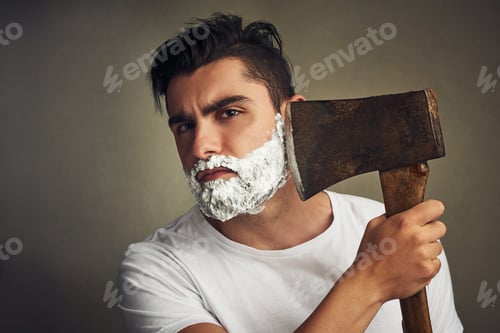 Preview: Its the right tool for the job. Shot of a handsome young man shaving his beard with a lumberjack.