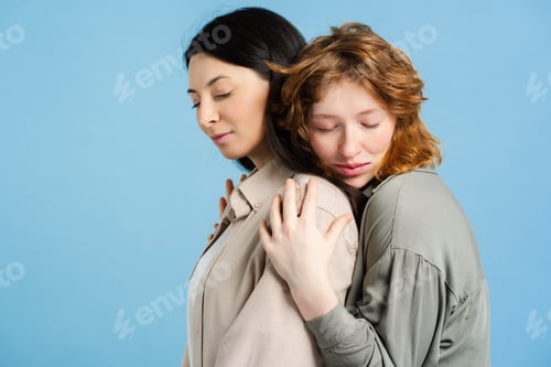 Preview: Portrait of beautiful pensive women hugging together with closed eyes isolated on blue background