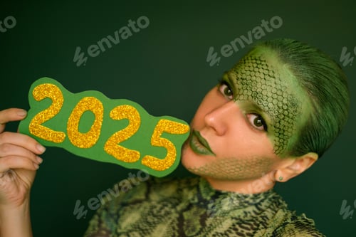 Preview: A woman with green makeup and painted snake scales on her face holds the numbers 2025.