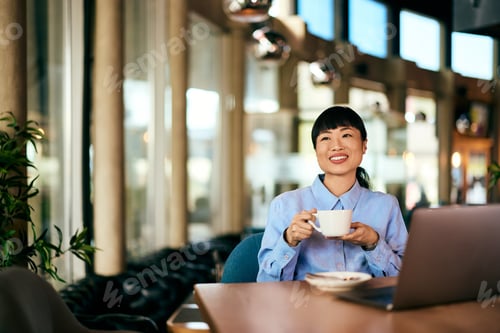 Preview: A smiling Asian woman, holding a cup of coffee, at the restaurant, working over the laptop.