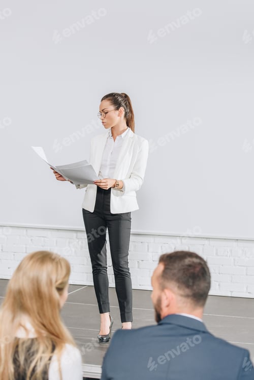 Preview: attractive business coach standing on stage and looking at documents during training in hub