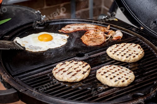 Preview: Burger patty, eggs and bacon grilling on barbecue grill plate . Backyard BBQ grill cooking burgers.