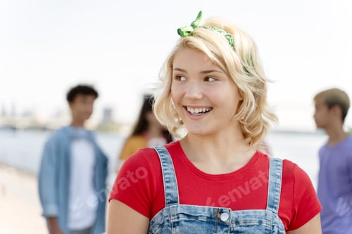 Preview: Smiling teenage girl wearing red t shirt looking at away on the street with friends on background