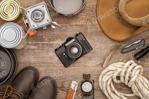 Preview: top view of photo camera, hat, boots and hiking equipment on wooden table