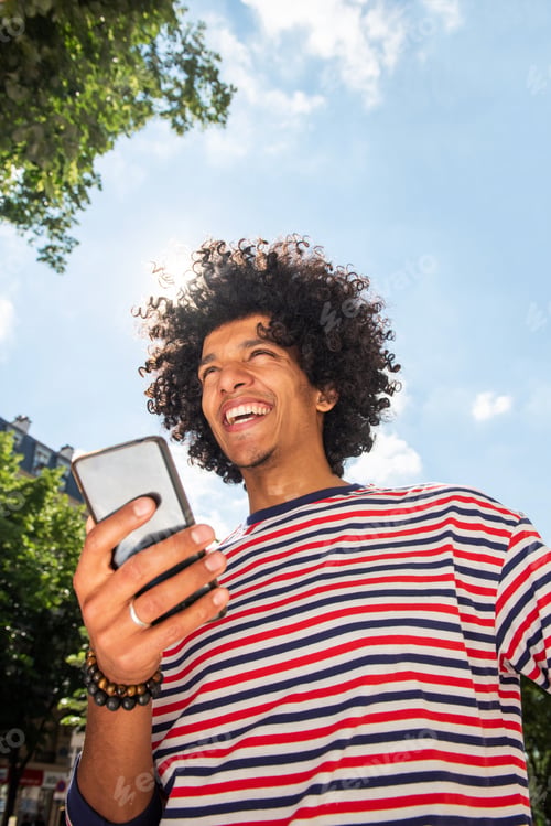 Preview: smiling young arabic man with afro holding mobile phone outside