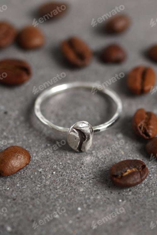 Preview: Elegant Silver Ring with Coffee Bean Detail Surrounded by Coffee Beans on Textured Surface.