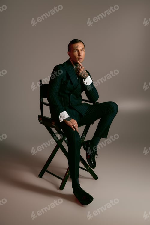 Preview: Handsome stylish man exuding charm in a sleek suit seated on a chair against a neutral background