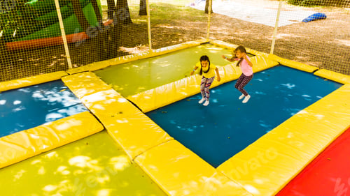 Preview: Little pretty girls having fun outdoor. Jumping on trampoline in children zone. Amusement park